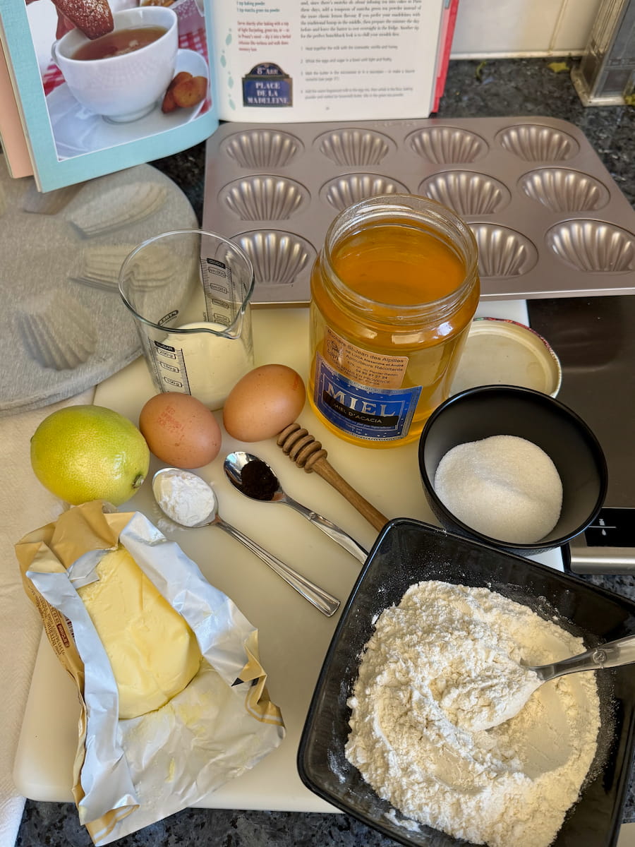 ingredients laid out to make madeleines cakes - flour, sugar, butter, eggs, vanilla, honey, baking powder and lemon