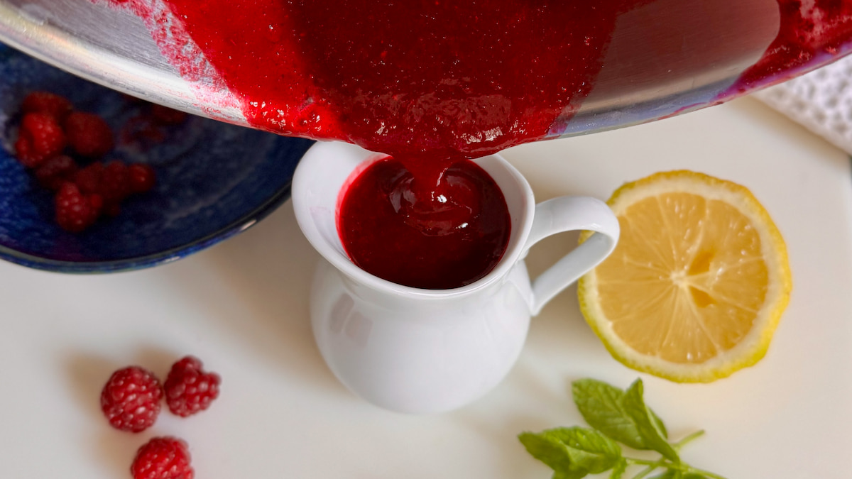 pouring a bright raspberry coulis into a little French porcelain jug next to lemon and fresh raspberries