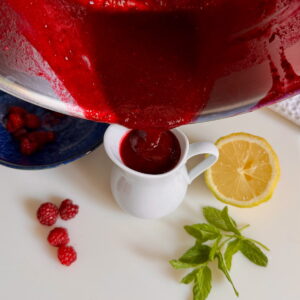 Pouring a raspberry coulis or sauce into a jug next to lemon and fresh raspberries