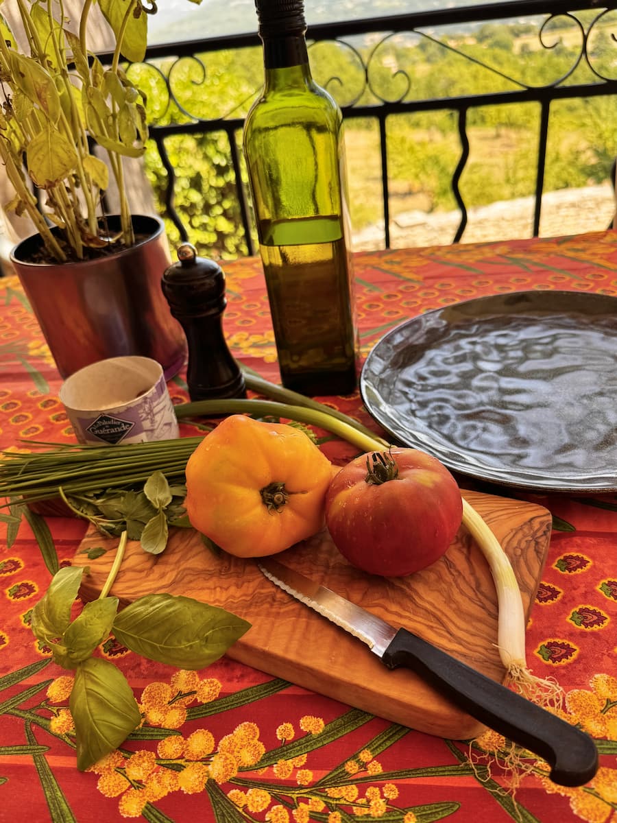 large red and yellow pineapple tomato on a chopping board on a Provence table, with olive oil, chives, basil, seasoning and a serrated knife