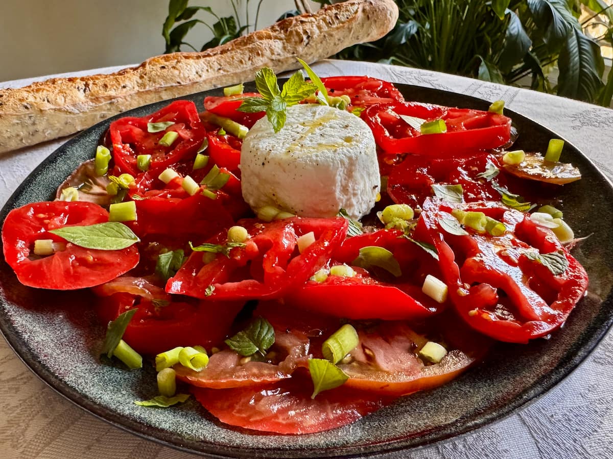 a ripe tomato salad served thinly sliced on a plate topped with chopped spring onions, fresh herbs and topped with fresh goat cheese with a French baguette