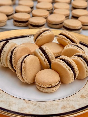plain ivory French macarons filled with a dark chocolate ganache on a large golden plate in front of a perfect batch of filled macarons